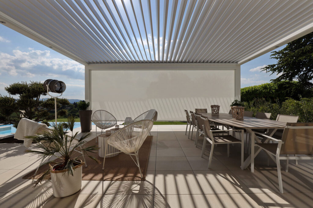 Pergola bioclimatique moderne avec lames orientables et store latéral, aménagée avec un salon extérieur et une grande table à manger, offrant un espace ombragé élégant avec vue sur la piscine et la nature.